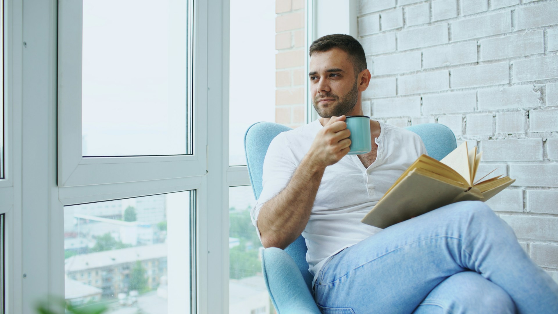 A man sitting by a window with coffee and a book, looking out thoughtfully